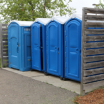 The Ultimate Guide to Portable Toilets: Your Solution for Outdoor Events and Construction Sites 3 Four blue portable toilets are lined up next to a wooden privacy fence outdoors, with a tree and parking lot visible in the background.