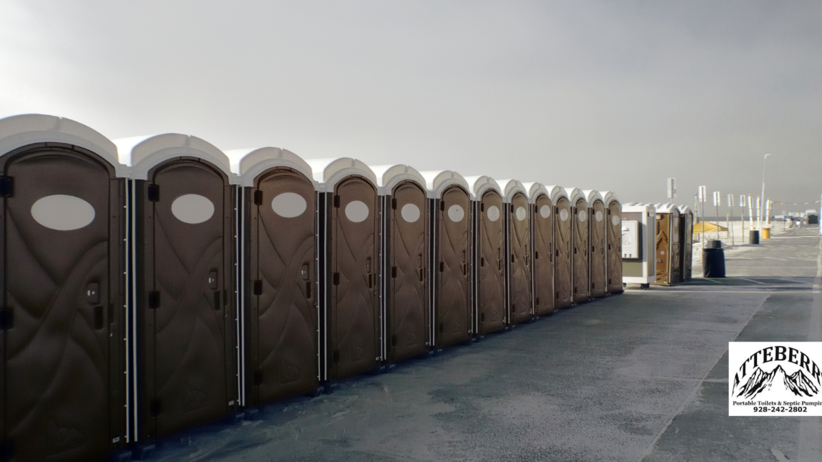 A row of portable toilets lined up on a paved outdoor area under a cloudy sky, with an Aztebert Portable Toilet Rentals sign in the foreground.