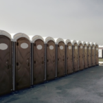 A row of portable toilets lined up on a paved outdoor area under a cloudy sky, with an Aztebert Portable Toilet Rentals sign in the foreground.