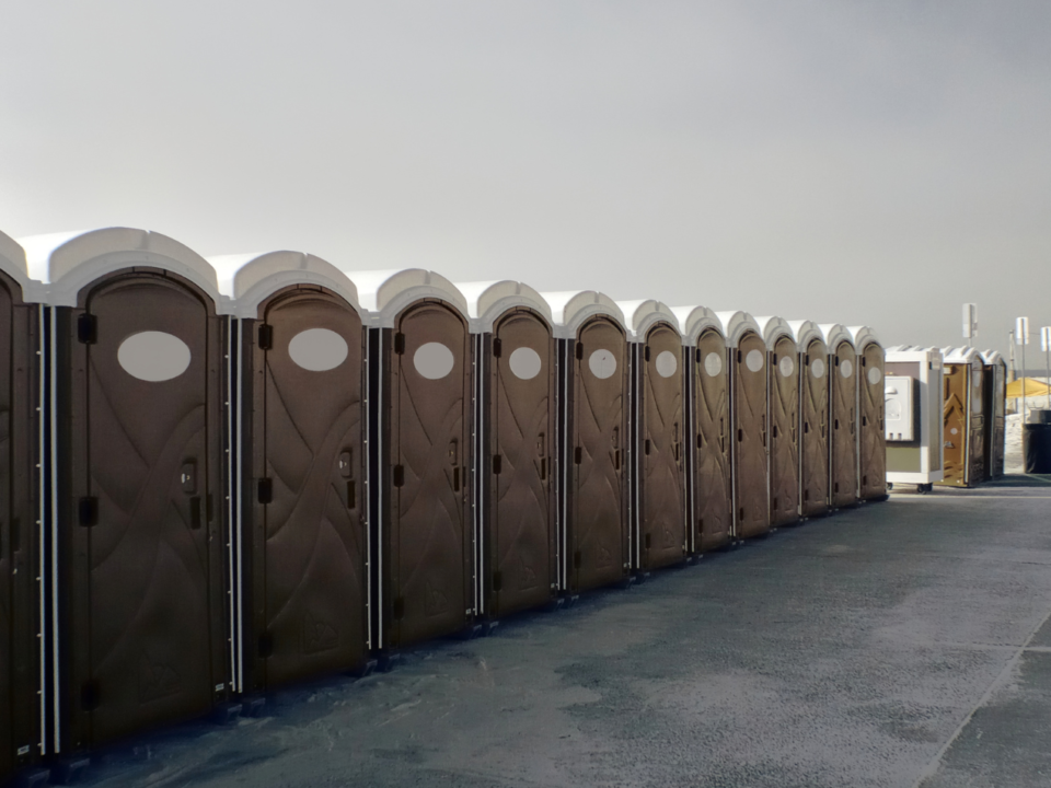 A row of portable toilets lined up on a paved outdoor area under a cloudy sky, with an Aztebert Portable Toilet Rentals sign in the foreground.