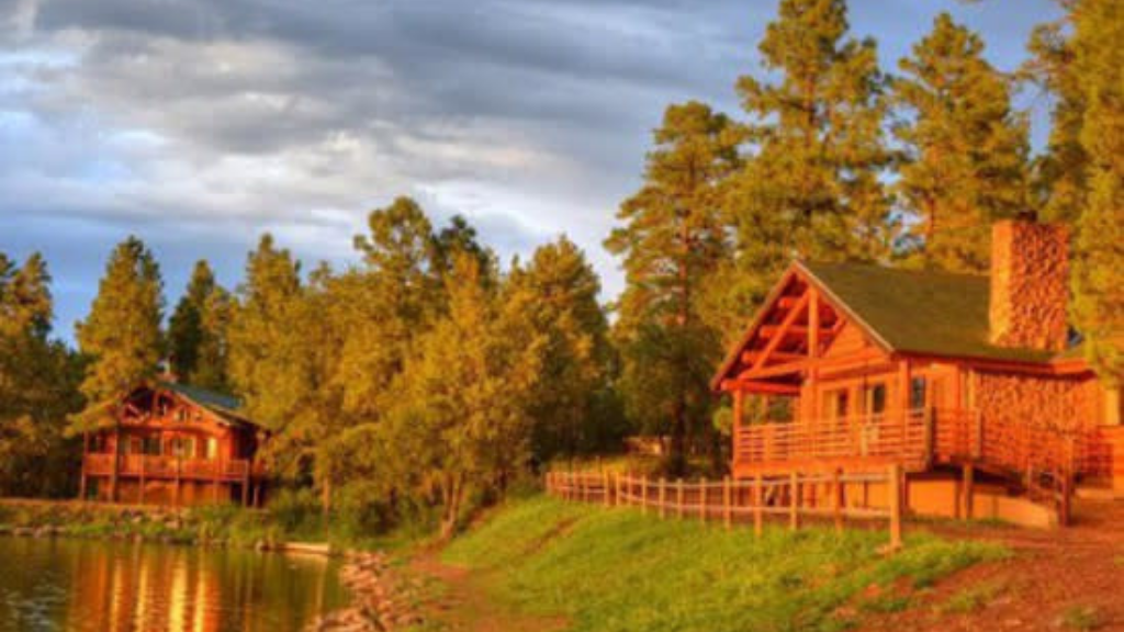 Two wooden cabins sit beside a calm lake, surrounded by trees and greenery, with warm sunlight casting a golden glow over the scene; a wooden fence lines the path near the water in Pinetop Lakeside, AZ.