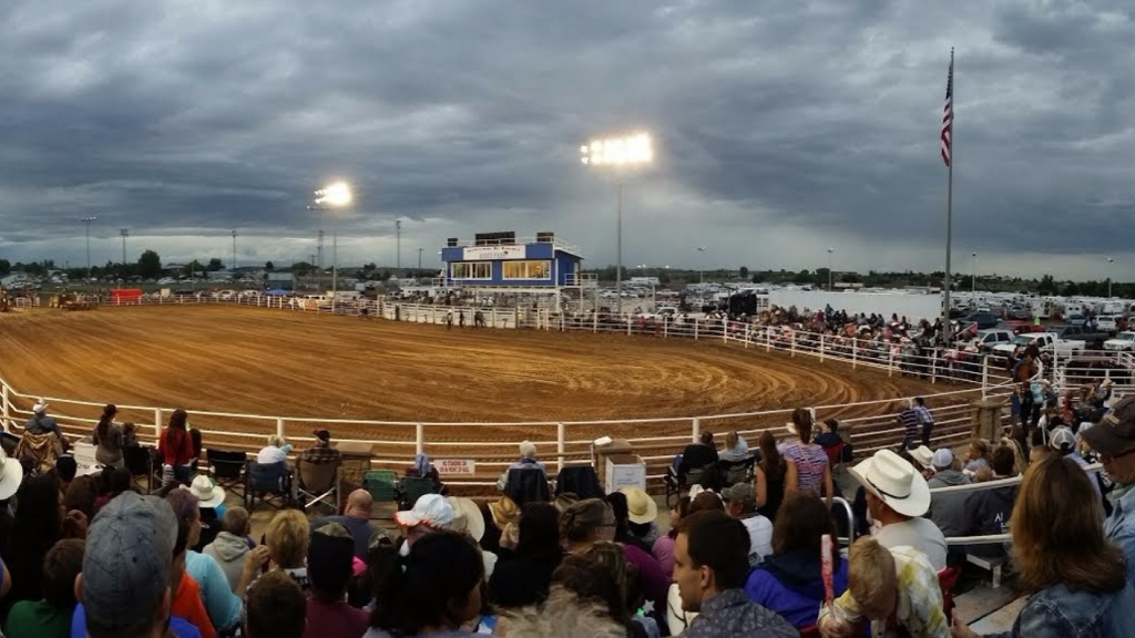 A large crowd watches an outdoor rodeo arena under cloudy skies, with stadium lights illuminating the dirt ring and an American flag flying on a tall pole. Spectators fill the stands, wearing hats and casual attire, in Taylor, AZ.