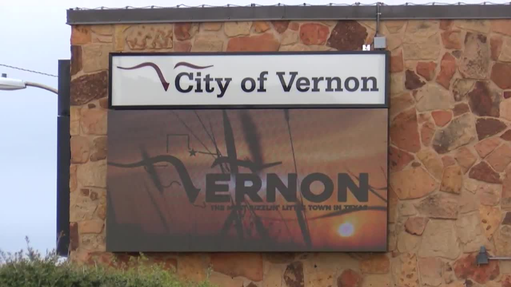 A sign on a stone wall reads City of Vernon. Below, a digital display features the word VERNON with a background of telephone poles against a sunset sky.